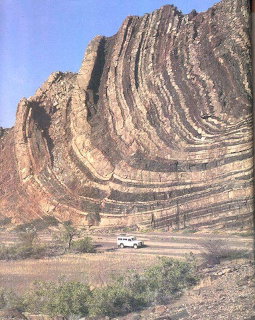 Layered folded rock cliff above a small white vehicle in a dry landscape