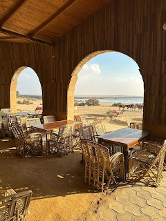 Open-air desert cafe with stone arches, wooden tables, wicker chairs, and a sandy landscape view