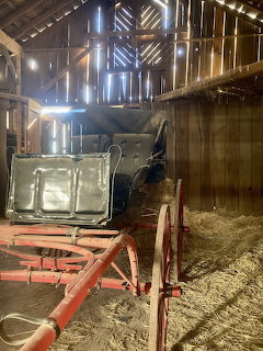 Old horse-drawn carriage inside a rustic wooden barn with hay on the floor and light through slats