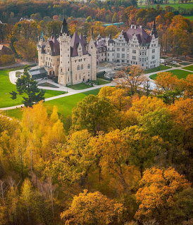 White castle with towers surrounded by colorful autumn trees and green lawns seen from above