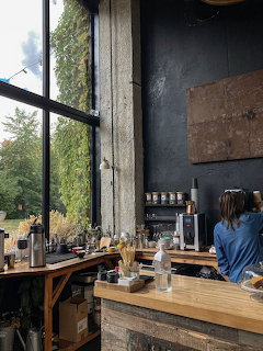 Interior of an industrial cafe with tall windows, wooden counter, and barista at the espresso machine