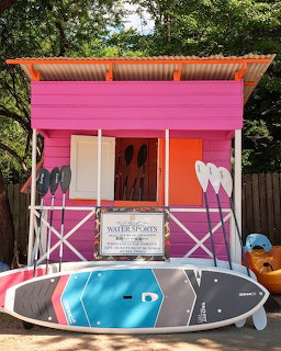 Bright pink surf rental shack with orange accents and paddleboard leaning against front fence