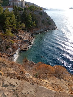 Rocky coastal cliff overlooking turquoise sea with hillside buildings and distant sailboat in Hydra, Greece
