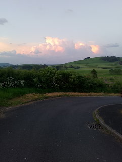 Curved country road with green hedgerows and rolling hills under pink sunset clouds