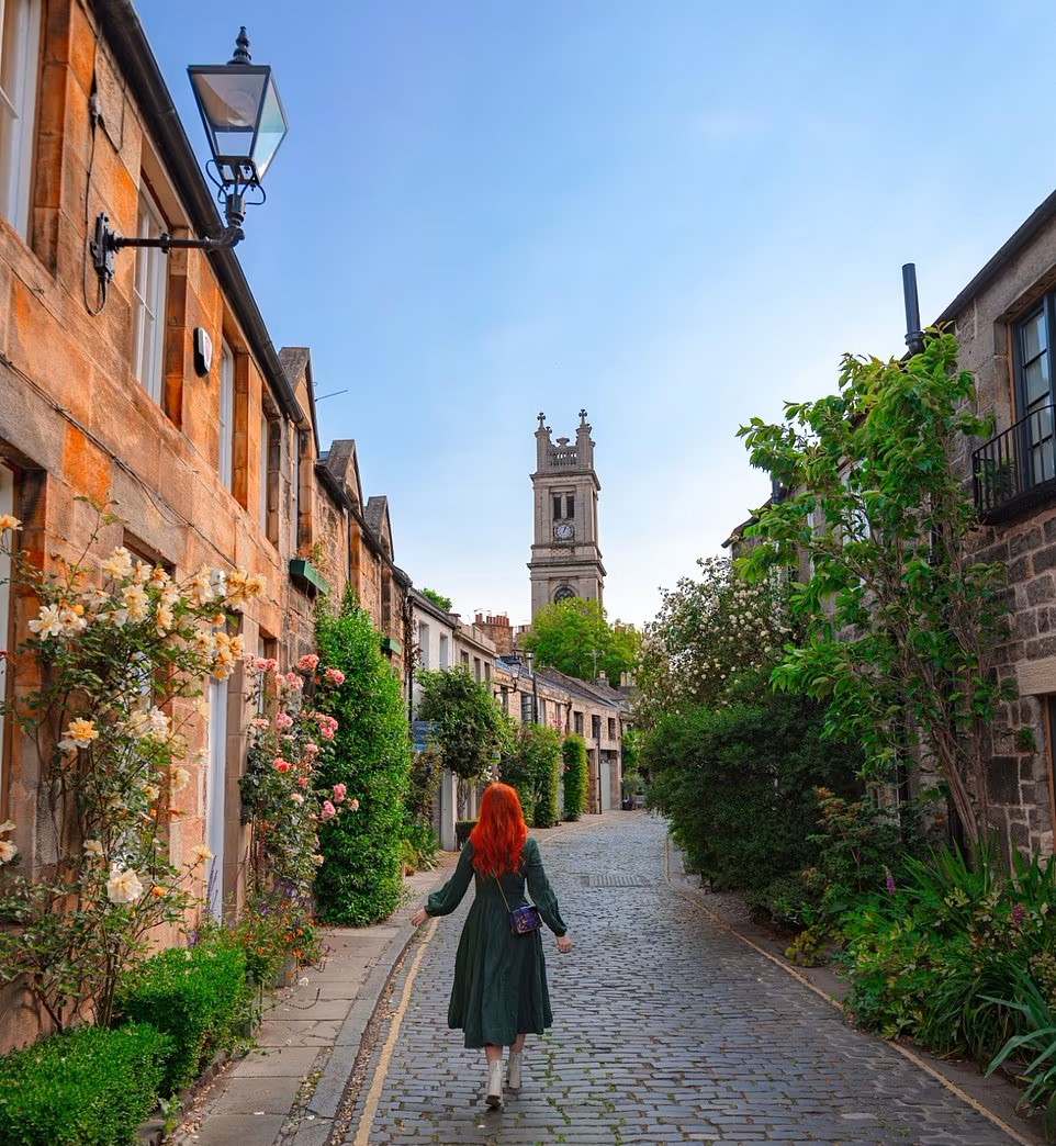 Woman walking down cobblestone lane lined with colorful buildings and greenery in Edinburgh, church tower visible ahead