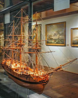 Detailed wooden tall ship model in a glass museum display case with maritime paintings on walls
