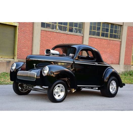 Black 1942 Willys coupe hot rod parked in front of a brick industrial building