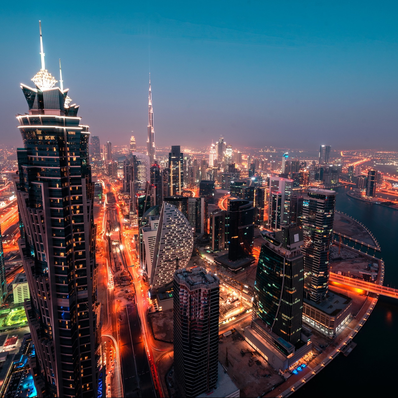 Nighttime Dubai skyline with Burj Khalifa, tall towers, bright roads, and a waterway