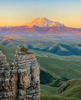 Snow-capped Mount Elbrus at sunset with green hills and rock formation in foreground