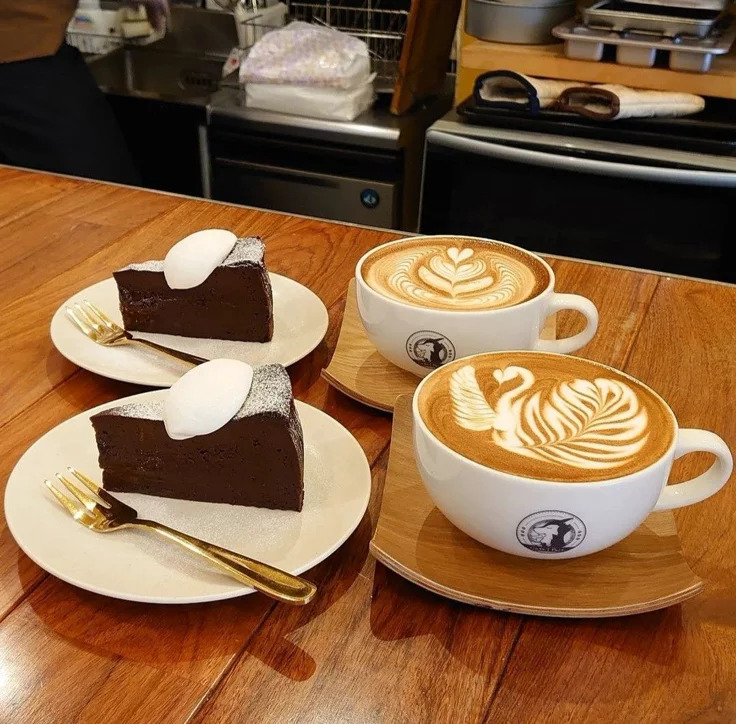 Two cappuccinos with latte art designs next to two slices of chocolate cake on white plates
