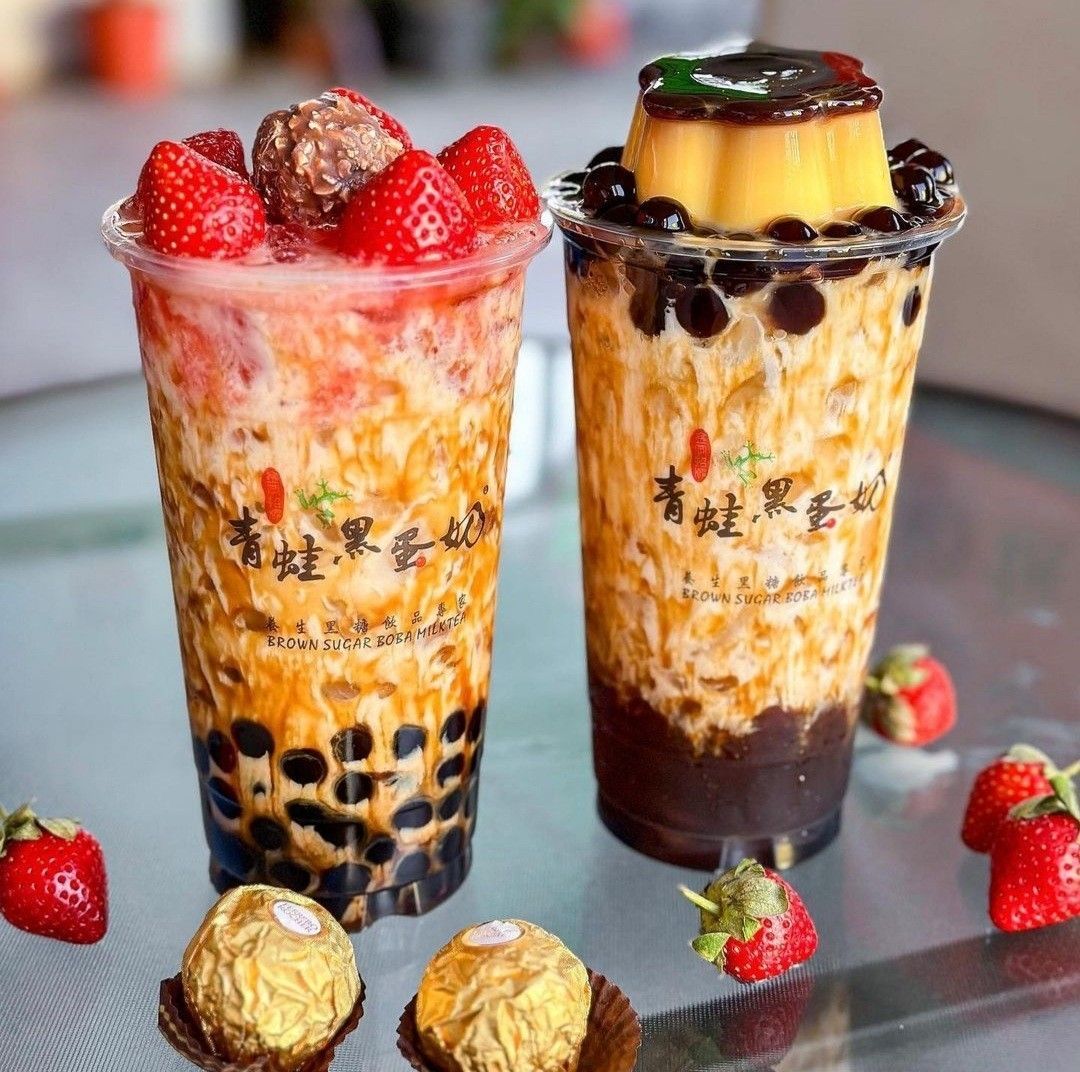 Two boba milk tea drinks with tapioca pearls, topped with strawberries and flan, strawberries and chocolates around them