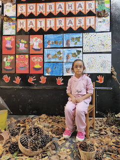 Young girl in pink outfit sitting on a chair with pine cones and autumn leaves, children's art on wall behind