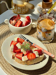Toast topped with strawberries and white cheese beside coffee and a bowl of fruit on a woven placemat