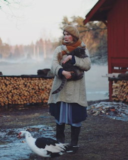 Woman in winter clothing holding a child outdoors near firewood and a red building