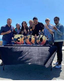 Seven people posing behind a table with a gold 300 number display outdoors near the ocean