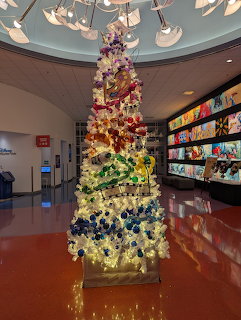 Tall Christmas tree with rainbow ombre decorations standing in a bright lobby with glossy floor and shelves behind