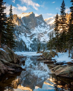 Snow-capped mountain peaks perfectly mirrored in a tranquil alpine lake surrounded by pine trees and rocks