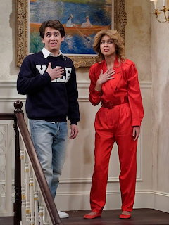 Woman in red suit and heels standing beside person in casual wear in elegant interior with framed painting