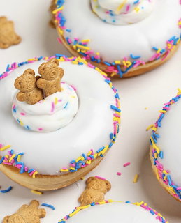 White-iced donuts decorated with colorful sprinkles and topped with tedy graham crackers and whipped cream