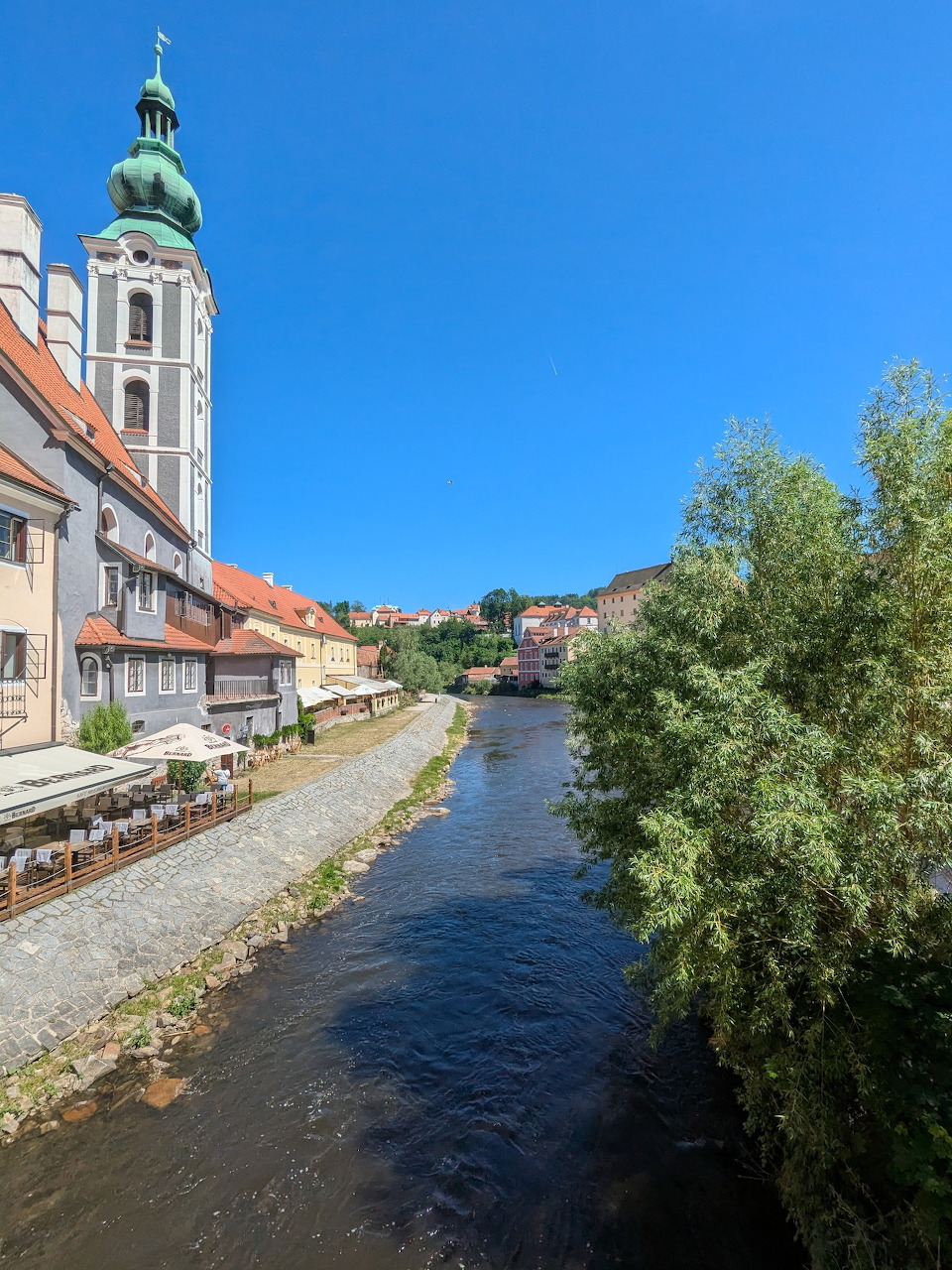 Historic town with a river, old buildings, a church tower, and a leafy tree under blue sky