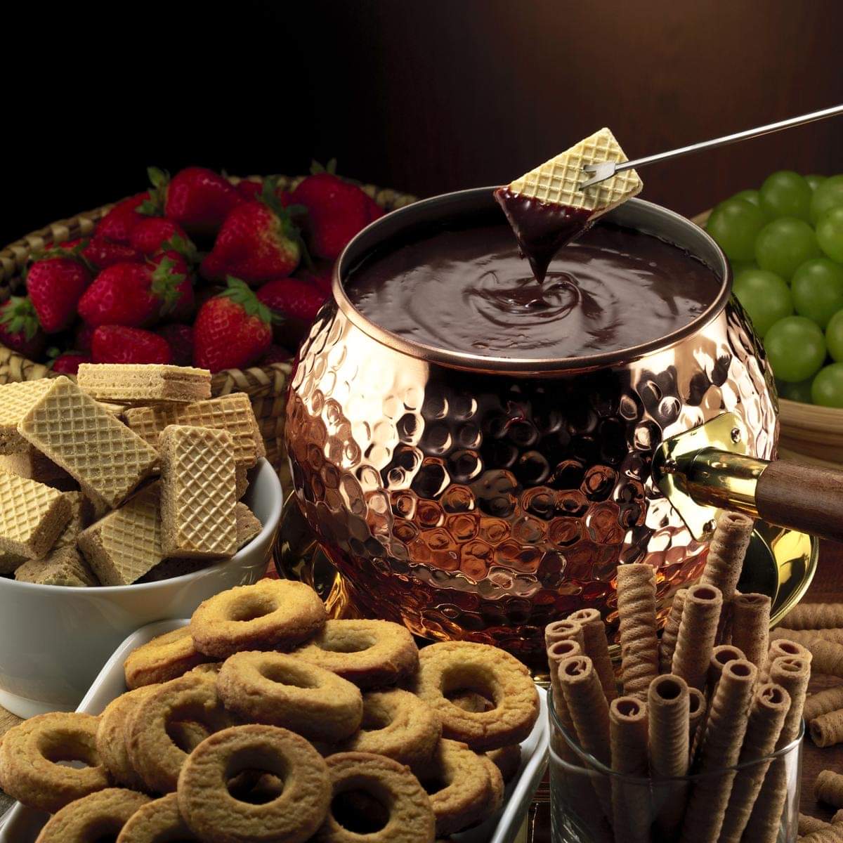Chocolate fondue in copper pot with strawberries, cookies, wafers, and pretzels arranged around it