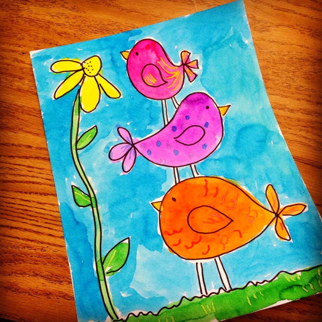 Childlike painting of colorful birds and flowers on blue paper on a wooden table