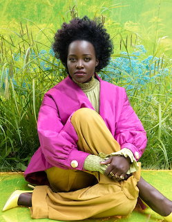 Lupita Nyong'o seated outdoors in a pink jacket and mustard yellow outfit amid grass and blue flowers