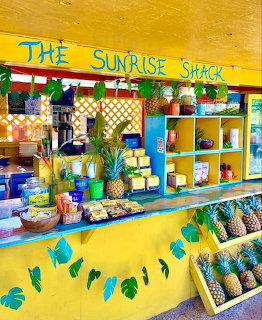 Bright yellow tropical shopfront with colorful shelves, plants, and pineapples at The Sunrise Shack