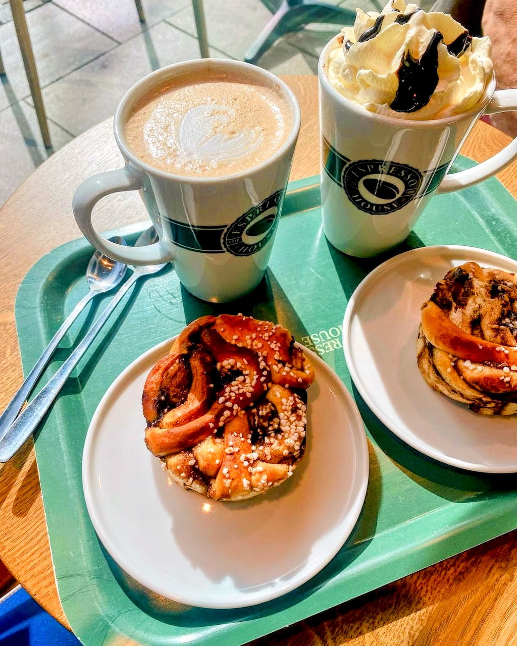 Two coffee drinks and pastries on plates arranged on a green tray on a wooden table