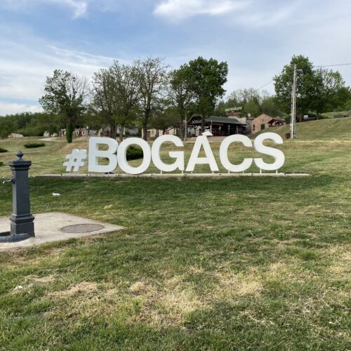 White BOGÁCS letters standing on green grass with trees and sky behind