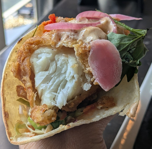 Close-up of a fish taco with fried white fish, pickled onions, greens, and sauce in a tortilla