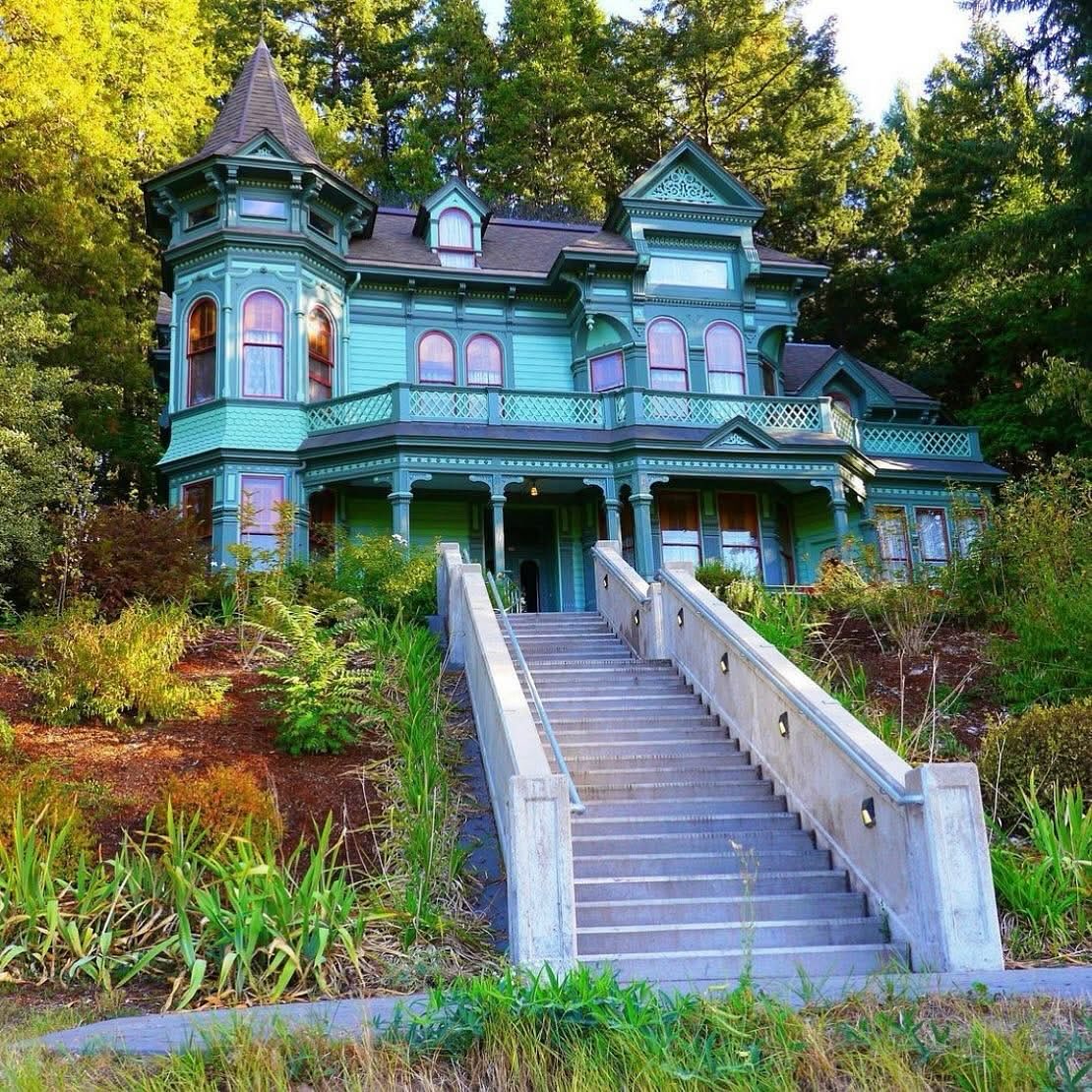 Turquoise Victorian house with pink trim, turrets, wrap-around porch, and white staircase surrounded by greenery
