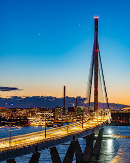 Illuminated cable-stayed bridge at sunset with city skyline and blue-orange sky