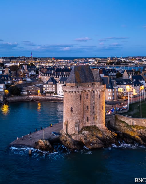 Stone tower on a rocky waterfront at dusk with harbor water and city buildings behind