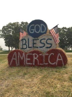 Hay bales painted with 'God Bless America' and two American flags in a grassy field.