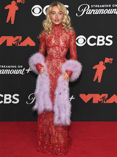 Woman in glamorous red sequined gown with white feather details posing on red carpet at MTV VMAs event