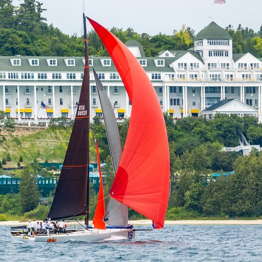 Sailing yacht with large red spinnaker sail racing past white waterfront hotel