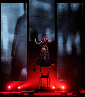Female performer on elevated stage platform with red lighting and large vertical video screens showing shadows