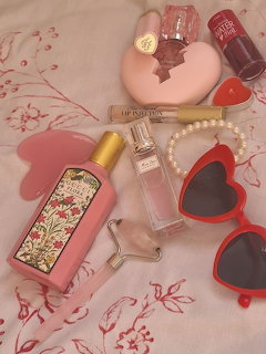Flat lay of pink beauty products including perfume, lipstick, heart sunglasses, pearls, and nail polish on floral fabric