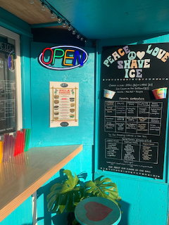 Turquoise shave ice stand interior with open neon sign, menu board, counter, and plants