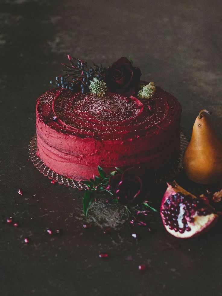 Red frosted cake with pomegranate seeds on decorative stand with pomegranate fruit and pear on dark background