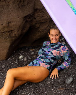 Woman in patterned wetsuit resting on black sand beach with surfboard