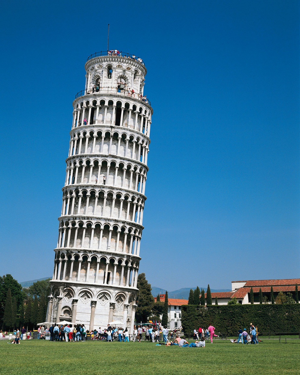 The Leaning Tower of Pisa tilting to the left with tourists on green grass and blue sky