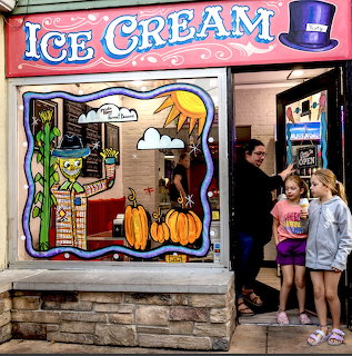 Two children viewing colorful ice cream shop window with painted decorations and pumpkins