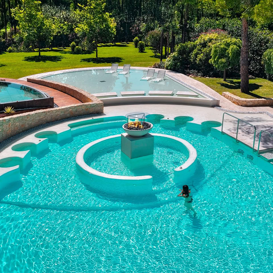 Aerial view of curved luxury pool with turquoise water and circular island surrounded by green landscape