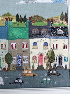 Textile art showing colorful patchwork houses on a village street with cars and trees made from various fabric pieces
