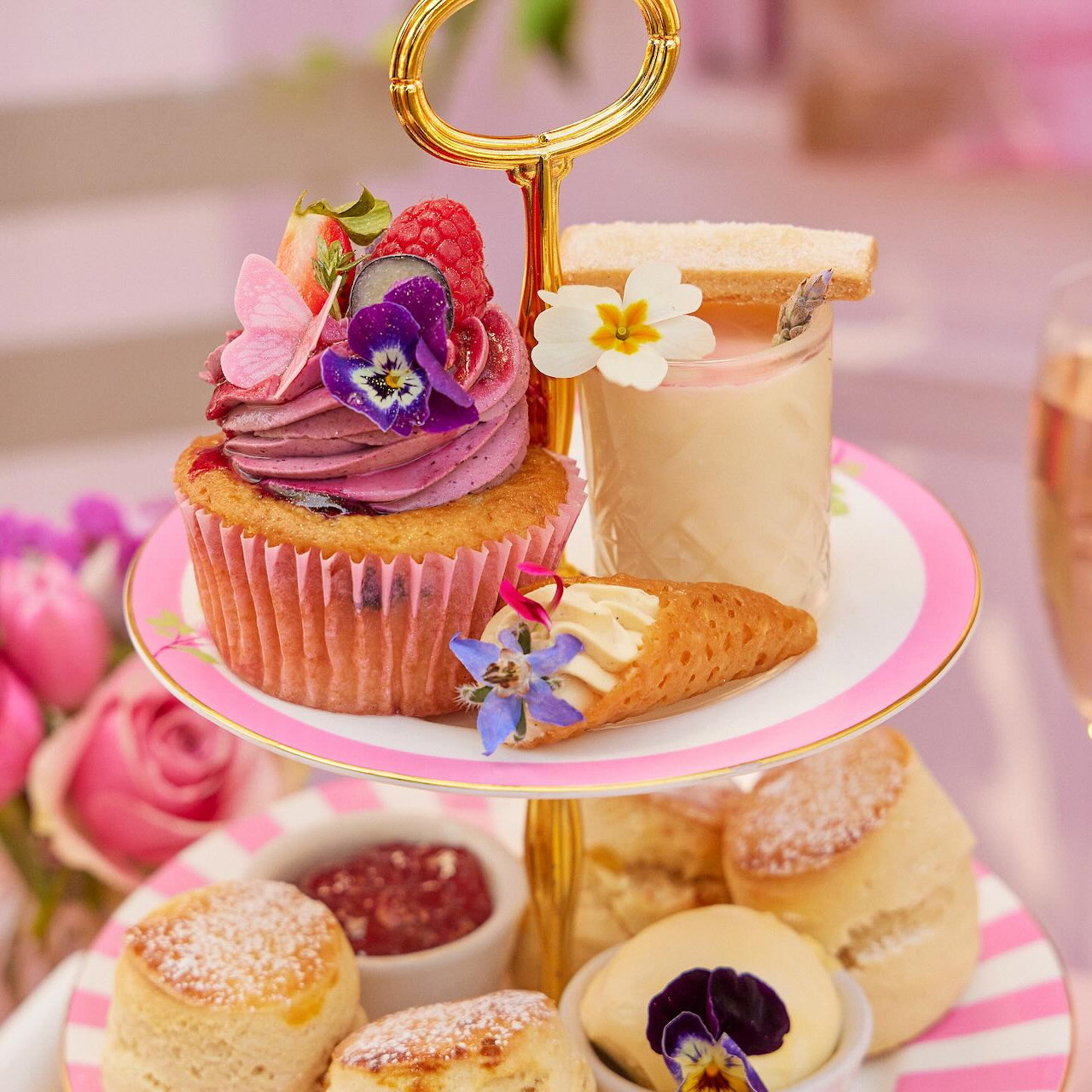 Three-tier gold stand with purple cupcake, pastries, cookies, and tea cup surrounded by pink flowers
