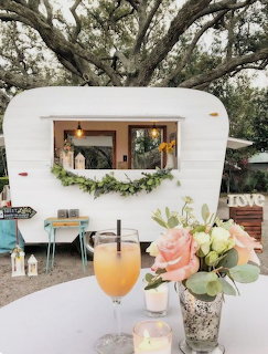 White vintage trailer bar with window serving area, fresh flowers, and orange drink on outdoor table