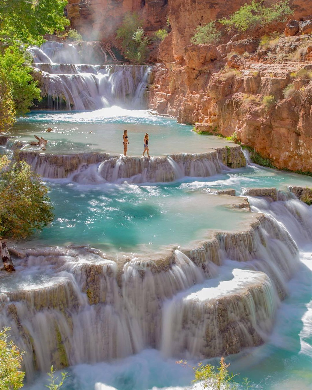 Cascading turquoise waterfalls flowing over white travertine terraces with two people standing in the pools