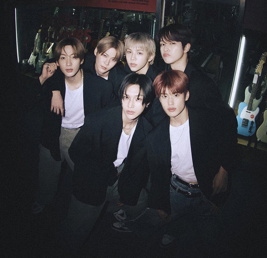 Six members of RIIZE kpop group posing together in dark suits against moody indoor backdrop with candles